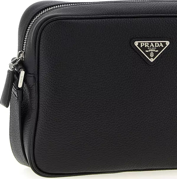 A close-up of a black Prada leather crossbody bag, showing the pebbled texture, silver zipper, and the iconic silver-tone triangular Prada logo plaque on the front.