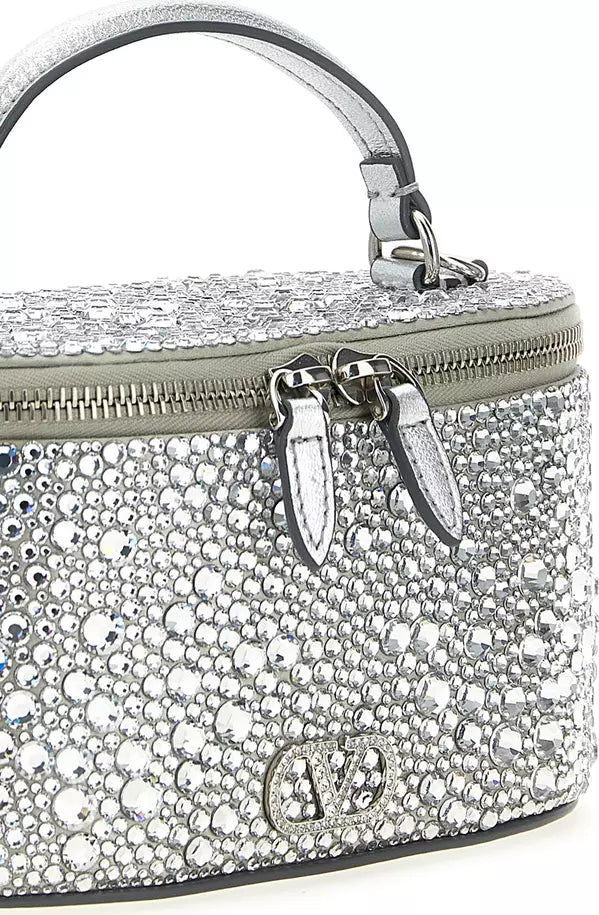 A silver rhinestone-covered top-handle handbag with a zippered top, silver-tone hardware, and a small oval logo plaque on the front, shown on a white background.