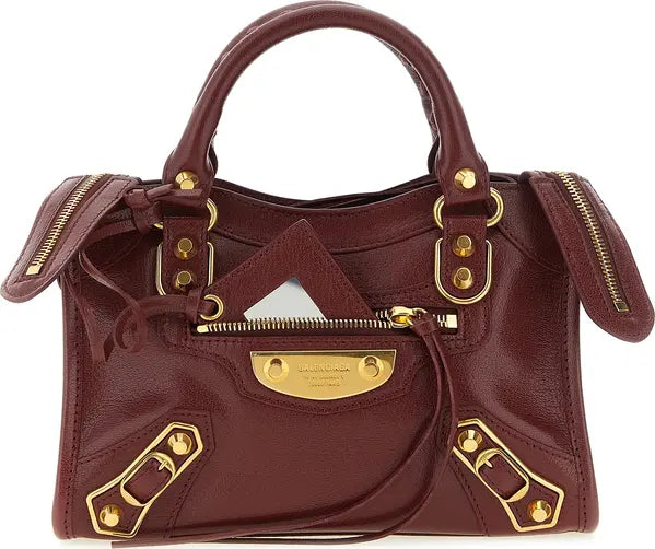 A dark burgundy leather handbag with gold-tone hardware, featuring a structured top handle, zippered front pocket, and decorative strap details, photographed against a plain white background.