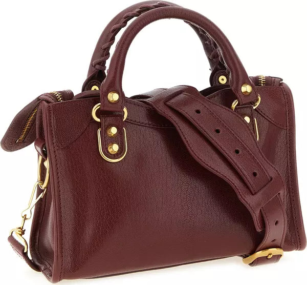 A burgundy leather handbag with gold-tone hardware, featuring a top handle, braided handle detail, a detachable shoulder strap, and a zippered side pocket, shown on a white background.