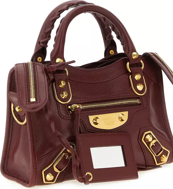 A dark burgundy leather handbag with gold-tone hardware, featuring braided top handles, multiple zippered compartments, and a front card holder with a blank white slot.