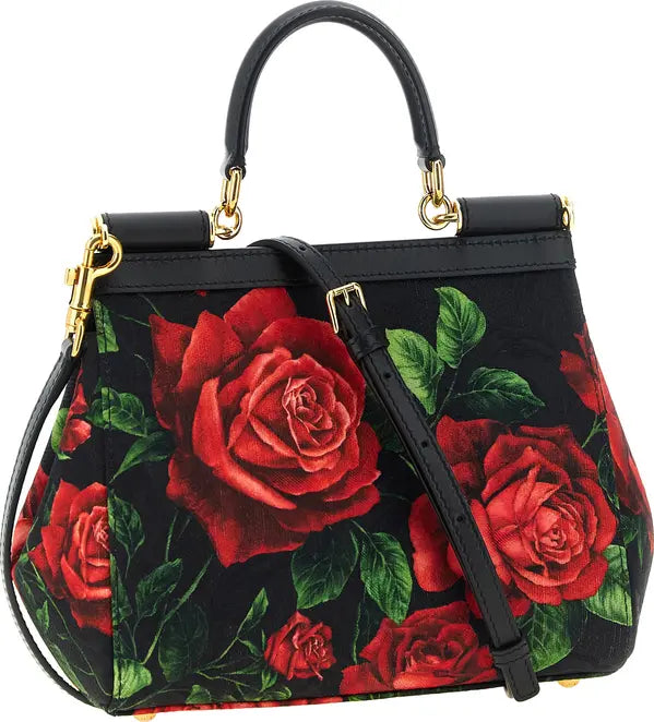 A structured black handbag with a large red rose and green leaf floral print, featuring black leather top handles, gold-tone hardware, and a detachable shoulder strap.