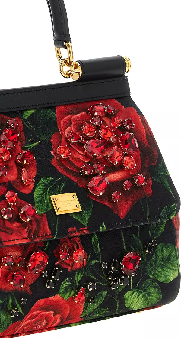 A close-up of a black handbag with a red rose print, embellished with large red crystal clusters and a gold-tone logo plaque.