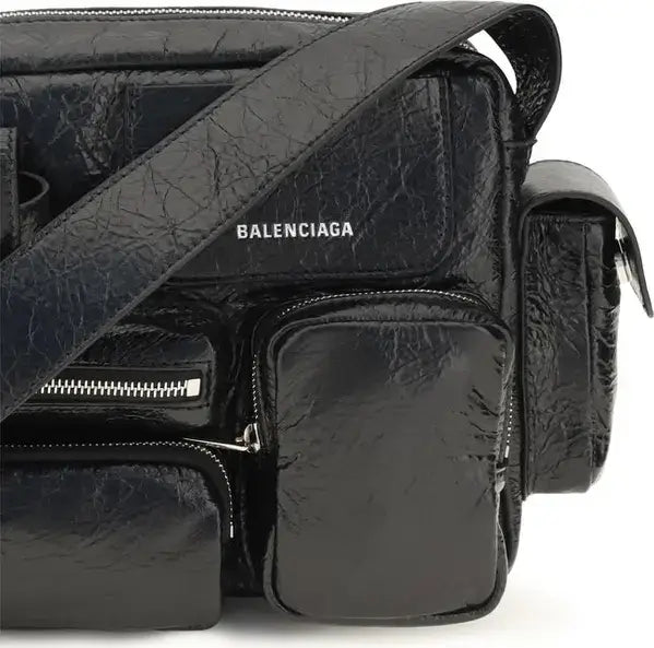 A close-up of a black Balenciaga shoulder bag with a crinkled textured finish, featuring multiple zippered pockets and a glossy patent leather side pouch. The Balenciaga logo is printed in white on the front.