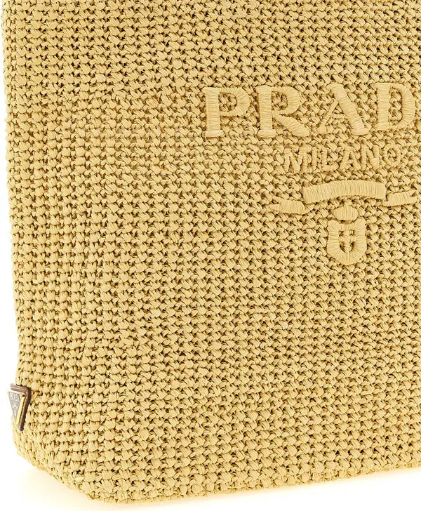 Close-up of a woven straw bag in a warm beige/tan color, featuring an embossed Prada Milano logo on the front.