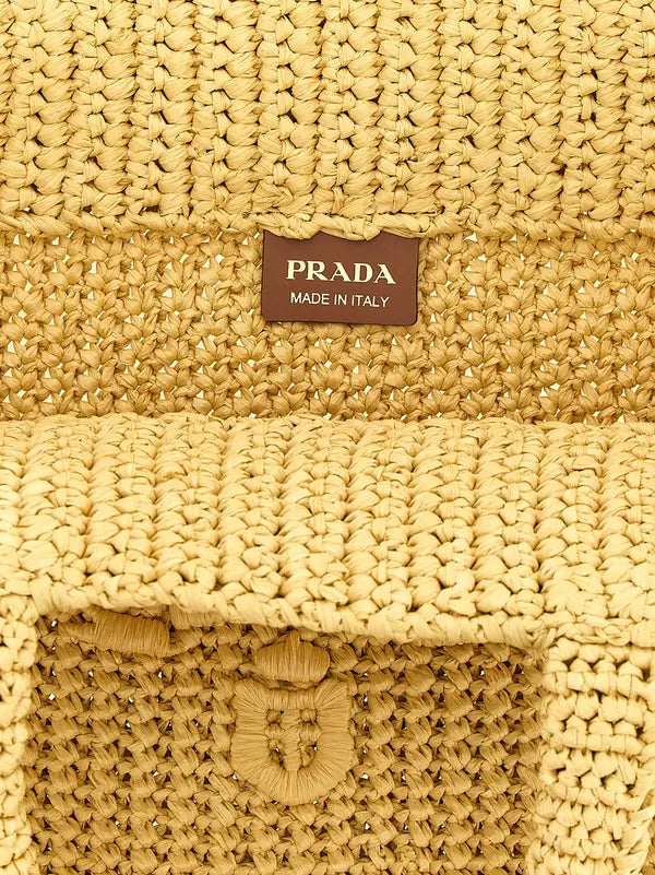 Close-up of a woven straw bag in a warm beige-yellow tone, featuring a brown rectangular Prada Made in Italy label stitched near the top center.