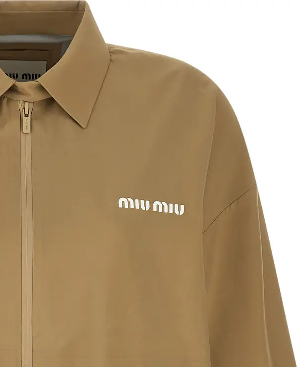 Close-up of a tan Miu Miu zip-up jacket with a pointed collar and white embroidered logo on the chest, set against a plain white background.