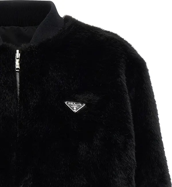 Close-up of a black fuzzy zip-up jacket with a silver zipper and a small silver Prada logo plaque on the chest, shown against a white background.