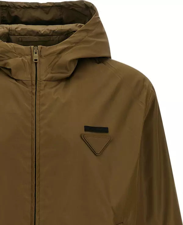 A close-up of a hooded jacket in a solid brownish-khaki color, featuring a front zipper, a zippered chest pocket with a triangular flap detail, and a black rectangular label, set against a plain white background.