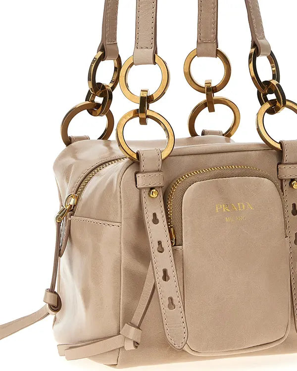 Prada taupe leather shoulder bag with gold-tone hardware, featuring a front zip pocket with gold-embossed logo, side pockets, and large interlocking ring handles on a white background.
