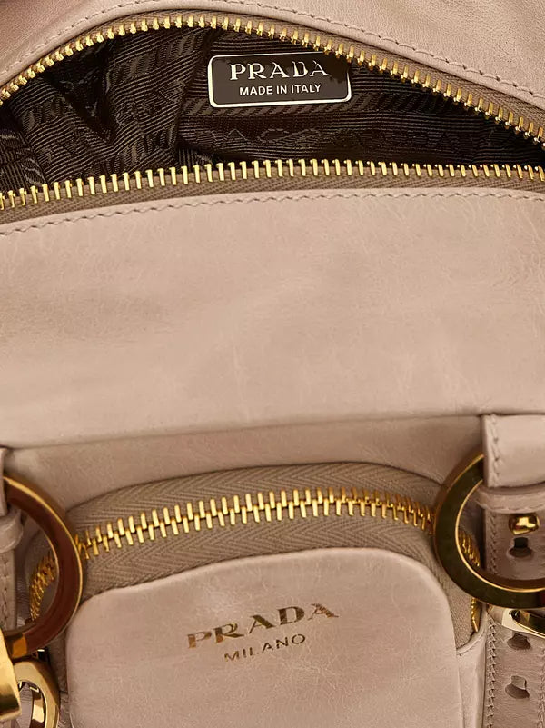 Close-up of a Prada handbag interior showing a light beige leather compartment with gold-tone zippers, a black patterned lining, and a Prada Made in Italy label.