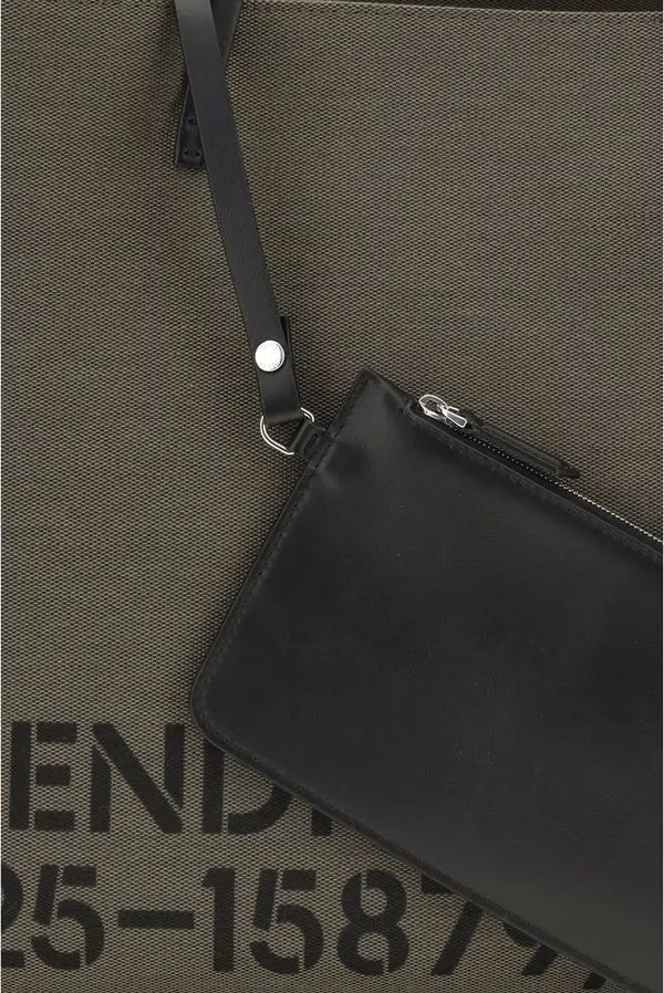 A close-up of a black leather wristlet pouch with a silver-tone metal snap and zipper, laid diagonally on a textured olive green fabric background with partial black lettering visible.