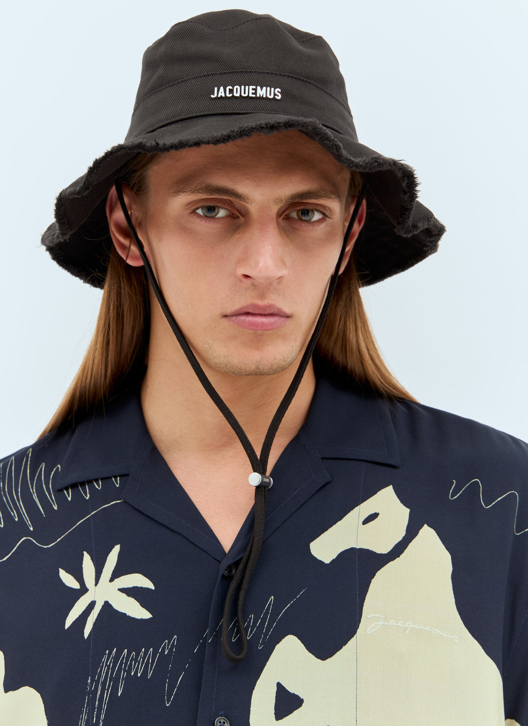 Man wearing a black bucket hat with 'Jacquemus' branding and a dark shirt with light designs.