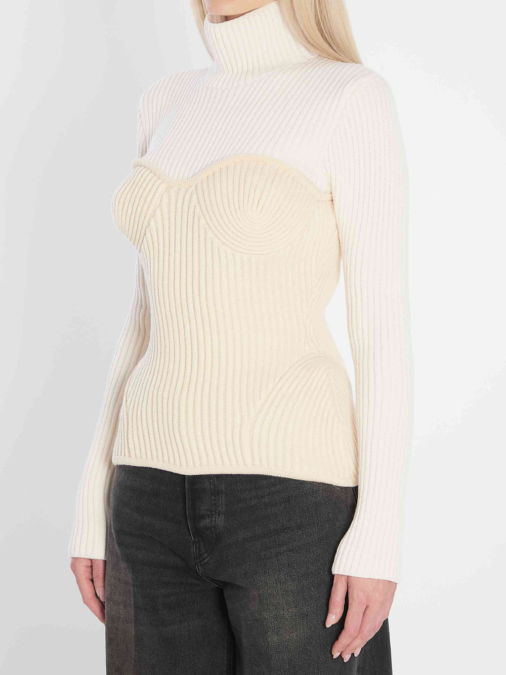 Woman wearing a cream ribbed sweater over a white turtleneck with dark jeans on a light gray background