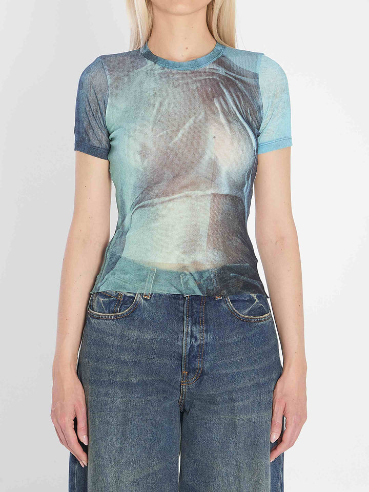 Person wearing a sheer blue top with a graphic design and jeans on a white background